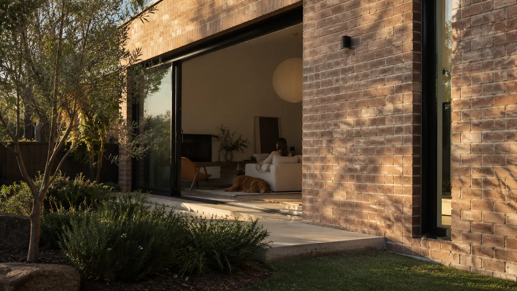 A brick house corner in warm evening light, with open glass doors showing a woman and dog relaxing in a bright living room
