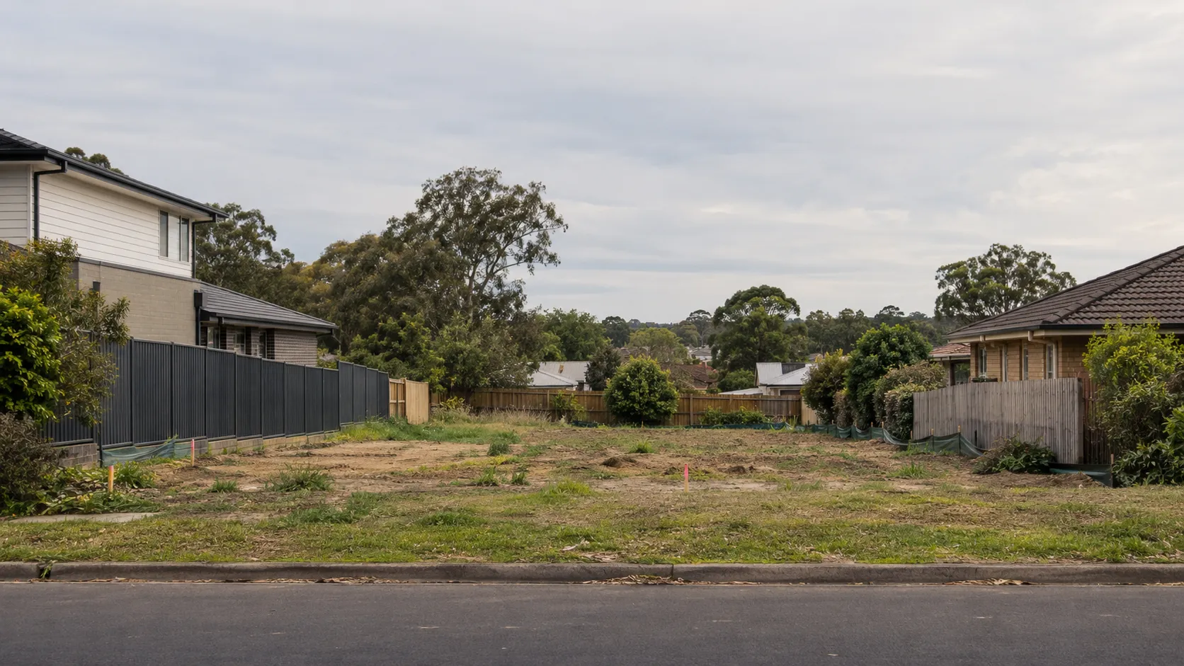 A vacant, grassed residential lot between two homes on a quiet street, with timber and metal fences and trees behind