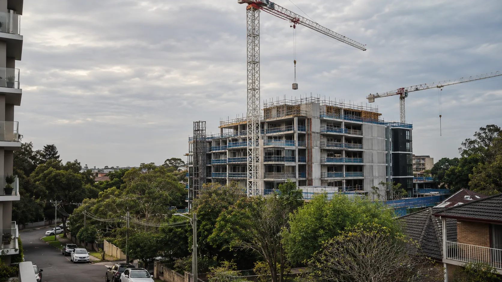 An apartment building under construction with scaffolding and two tower cranes above a leafy residential street