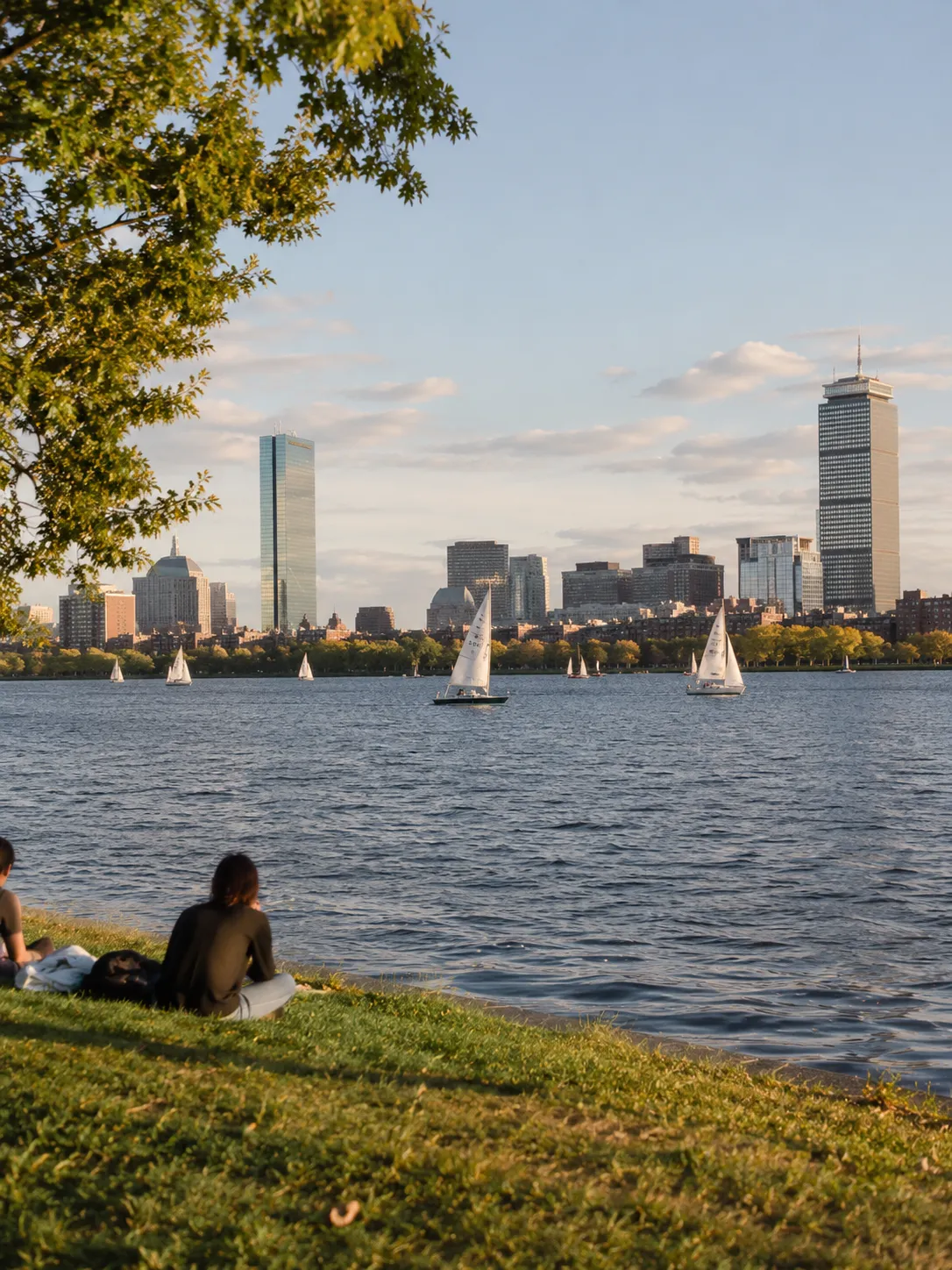 Boston skyline across the Charles River at sunset, with sailboats on the water and two people sitting on the grassy bank