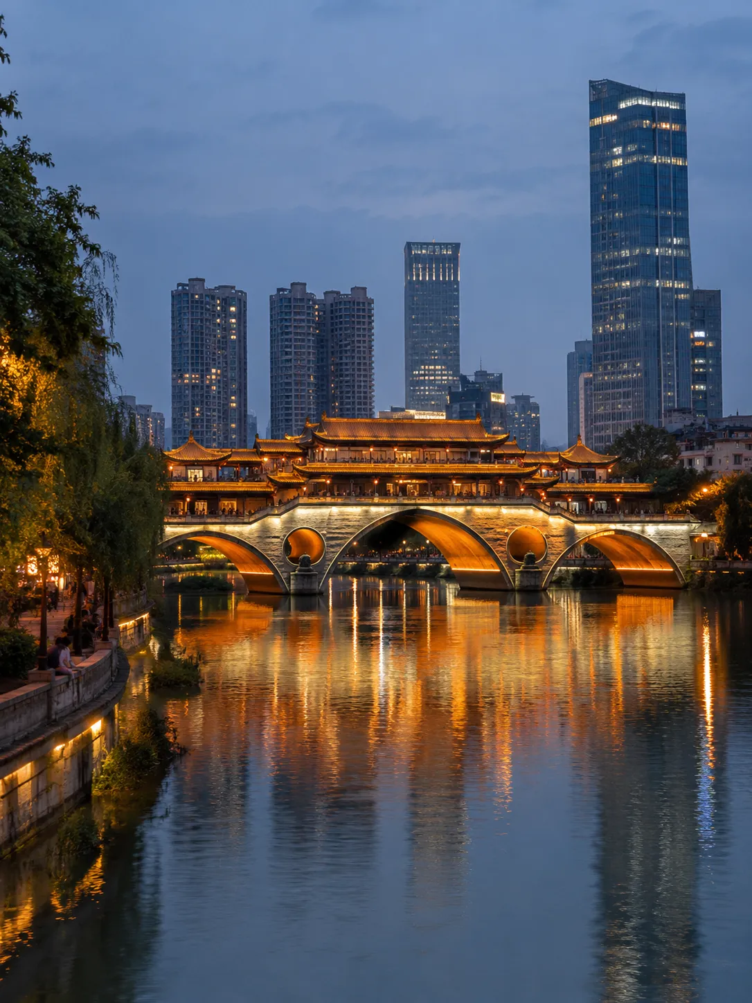 Chengdu at dusk, with an illuminated traditional covered bridge reflected in the river and modern skyscrapers rising behind it