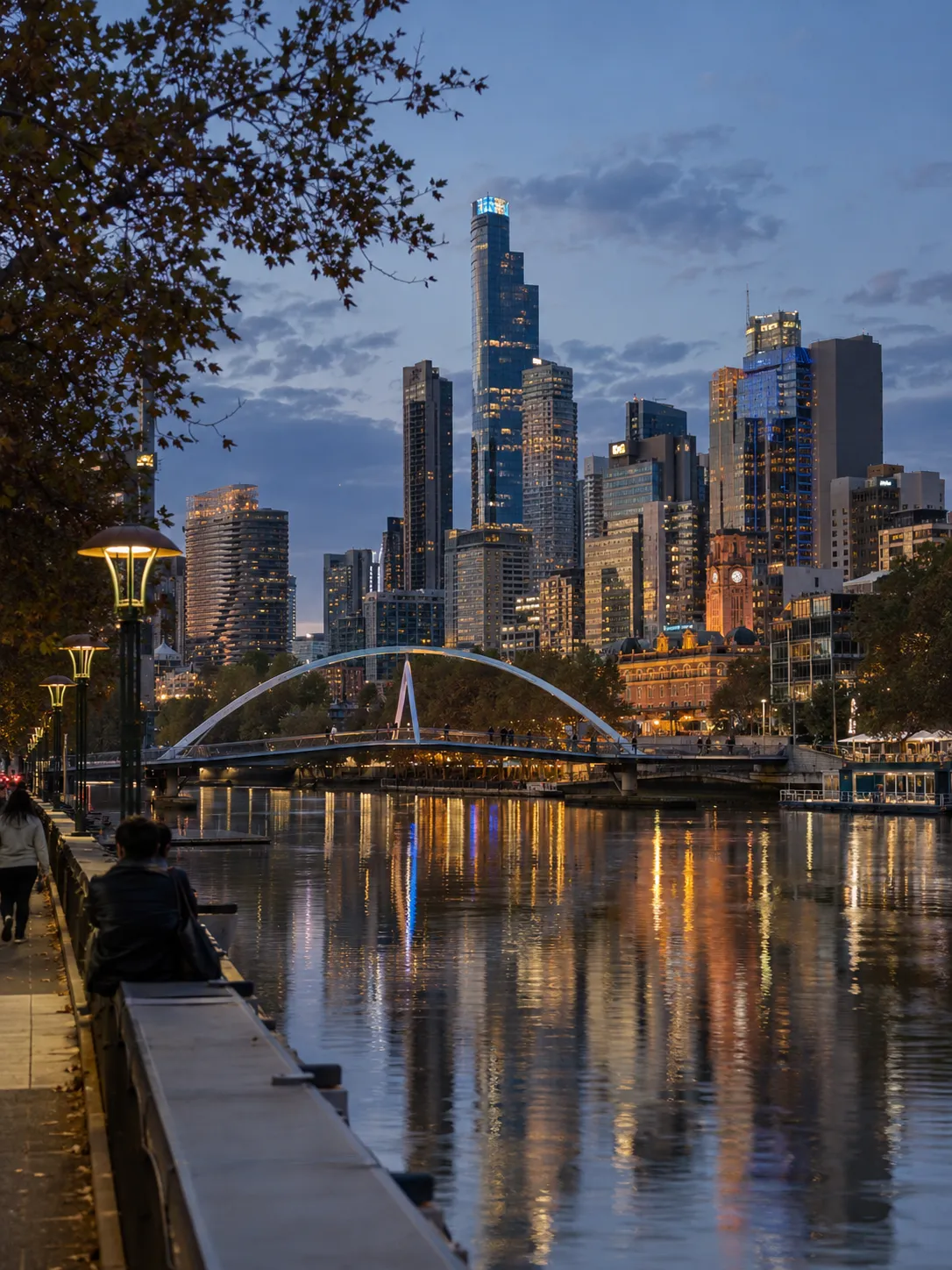 Melbourne skyline at dusk across the Yarra River, with a white arched footbridge and city lights reflected in the water