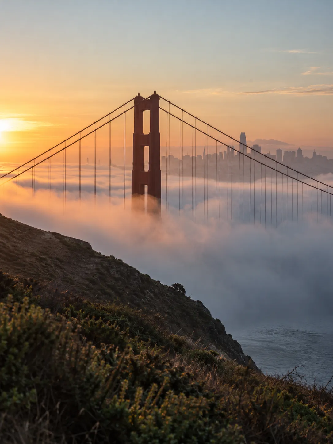 Golden Gate Bridge tower rising through low fog at sunrise, with the San Francisco skyline in the distance