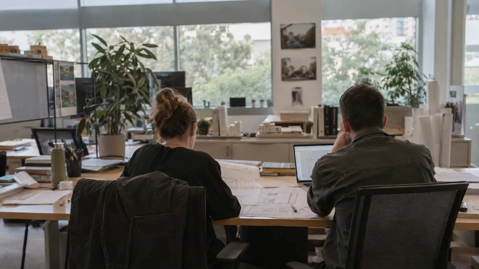 Two people at desks reviewing drawings and laptops in a bright open-plan office