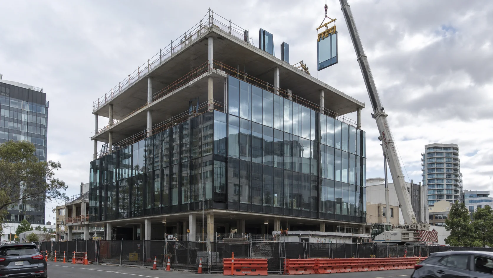 A crane lifting a glass panel onto a mid-rise building under construction with a glass curtain-wall façade