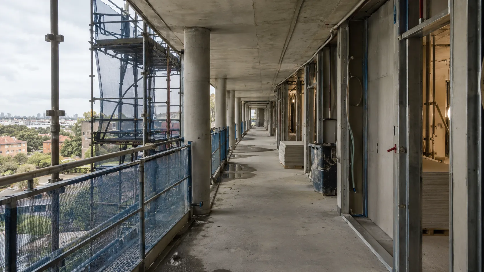 An unfinished apartment building walkway with a concrete slab floor, exposed columns, plasterboard framing, scaffolding and blue safety netting beside it