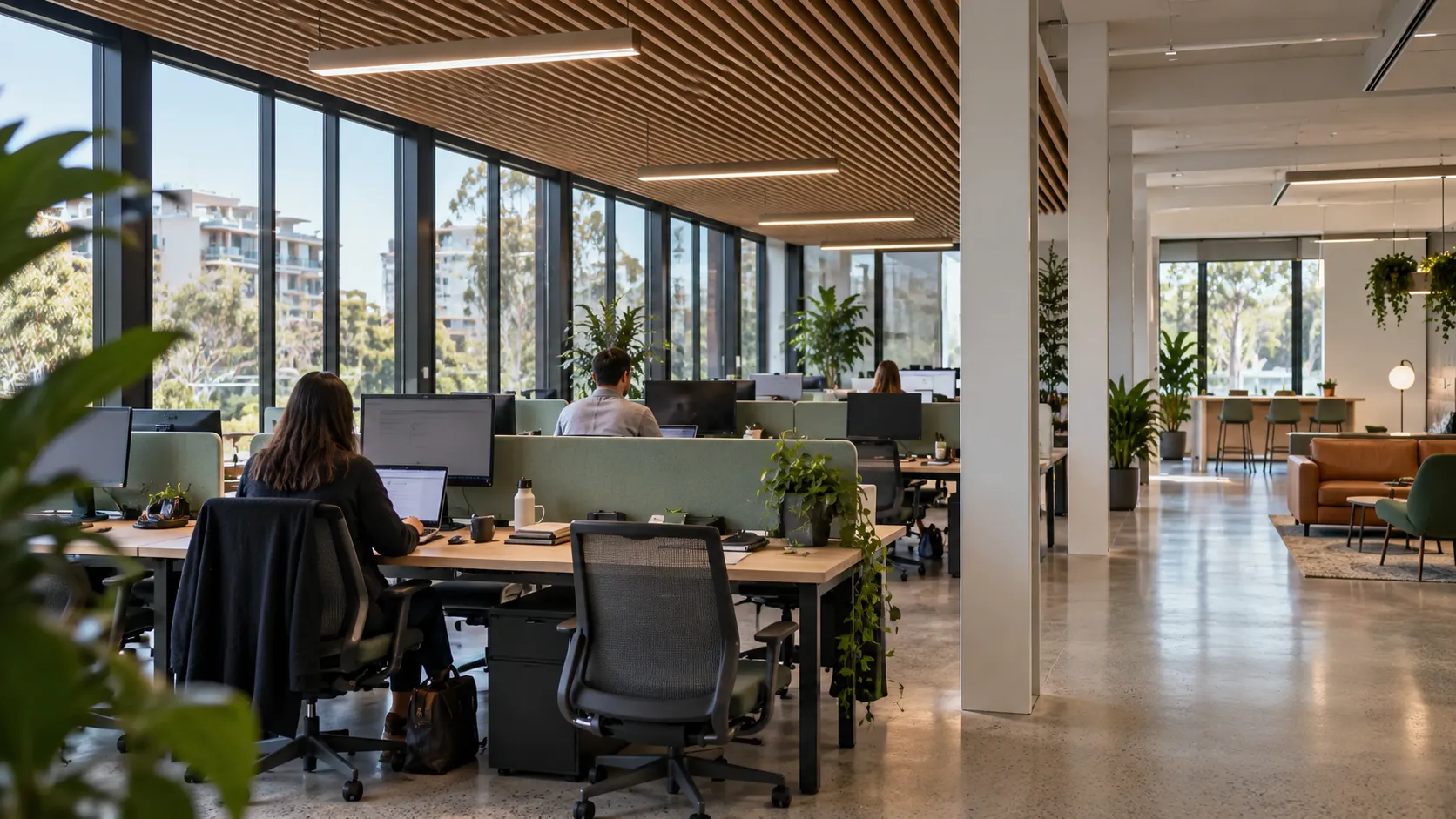 An open-plan office with timber slat ceiling, staff at desks with green screens, plants, polished concrete floor and a lounge area beyond