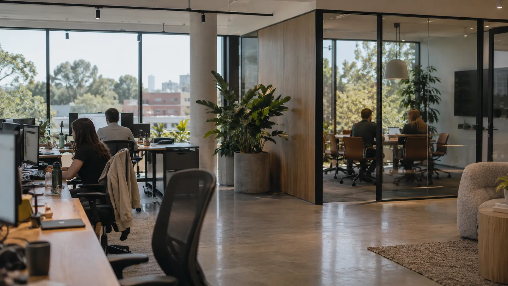 An office with a woman working at a desk in the foreground, a large plant, and two people meeting in a glass-walled room