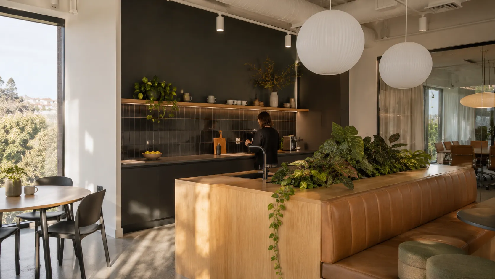 An office kitchen with a dark tiled wall and shelf, a person making coffee, a timber island with plants, a tan leather banquette and globe pendant lights