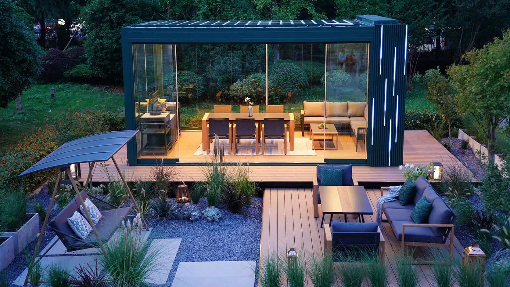 Glass-walled outdoor pavilion lit at dusk, with dining and lounge furniture on a timber deck