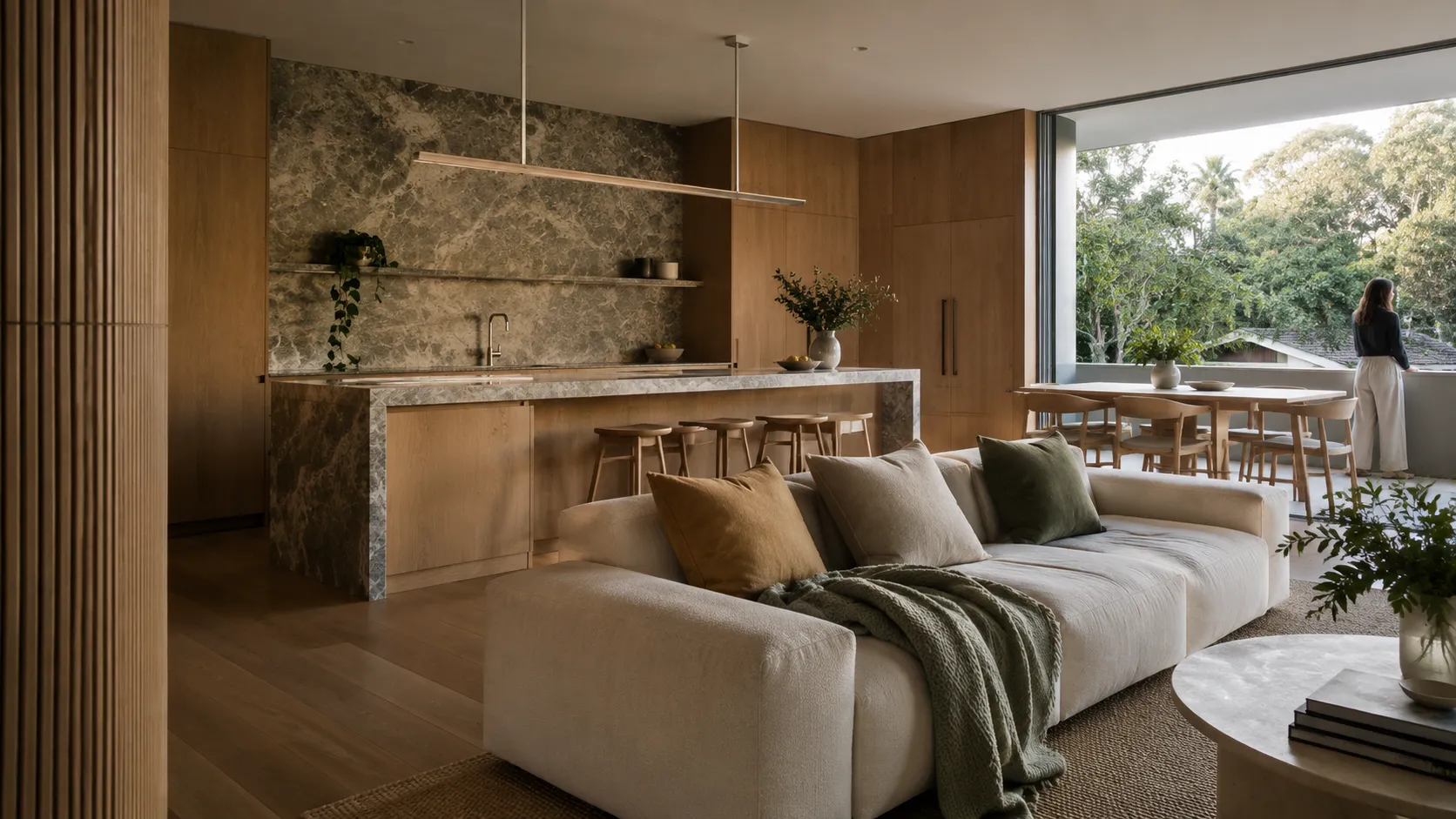 An open living space with a green-grey stone kitchen island and splashback, timber joinery, a cream sofa, a dining table and a balcony view of trees