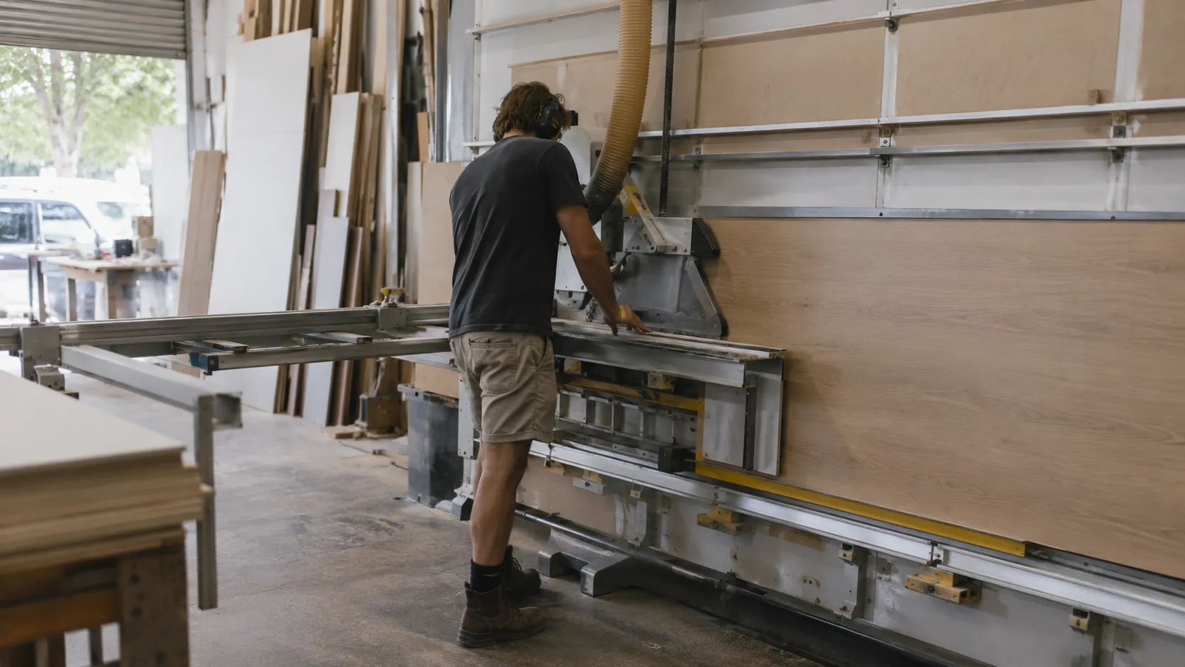 A workshop with a person wearing ear protection feeding a large timber board into a vertical panel saw, with stacked sheets behind
