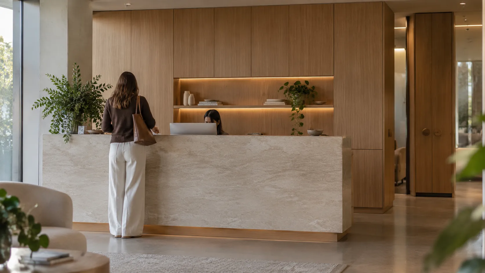 A reception counter clad in pale stone with timber joinery and lit shelving behind, a receptionist at a laptop and a visitor standing at the desk