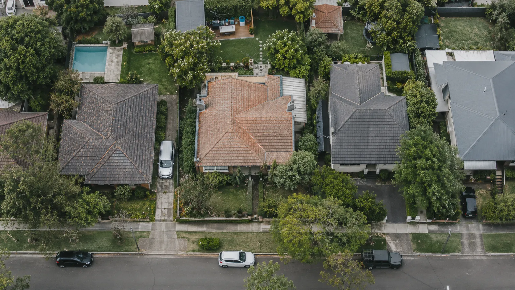 An aerial view of a suburban street with a terracotta-roofed house at the centre, neighbouring grey roofs, gardens, a pool and parked cars