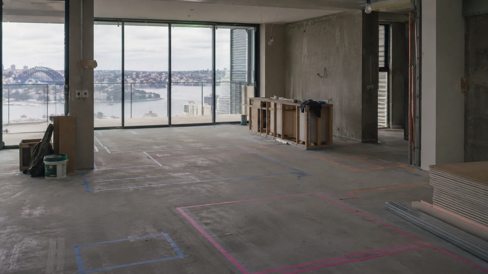 An unfinished high-rise interior with a concrete floor marked by coloured tape lines, a view of Sydney Harbour, building materials and a workbench