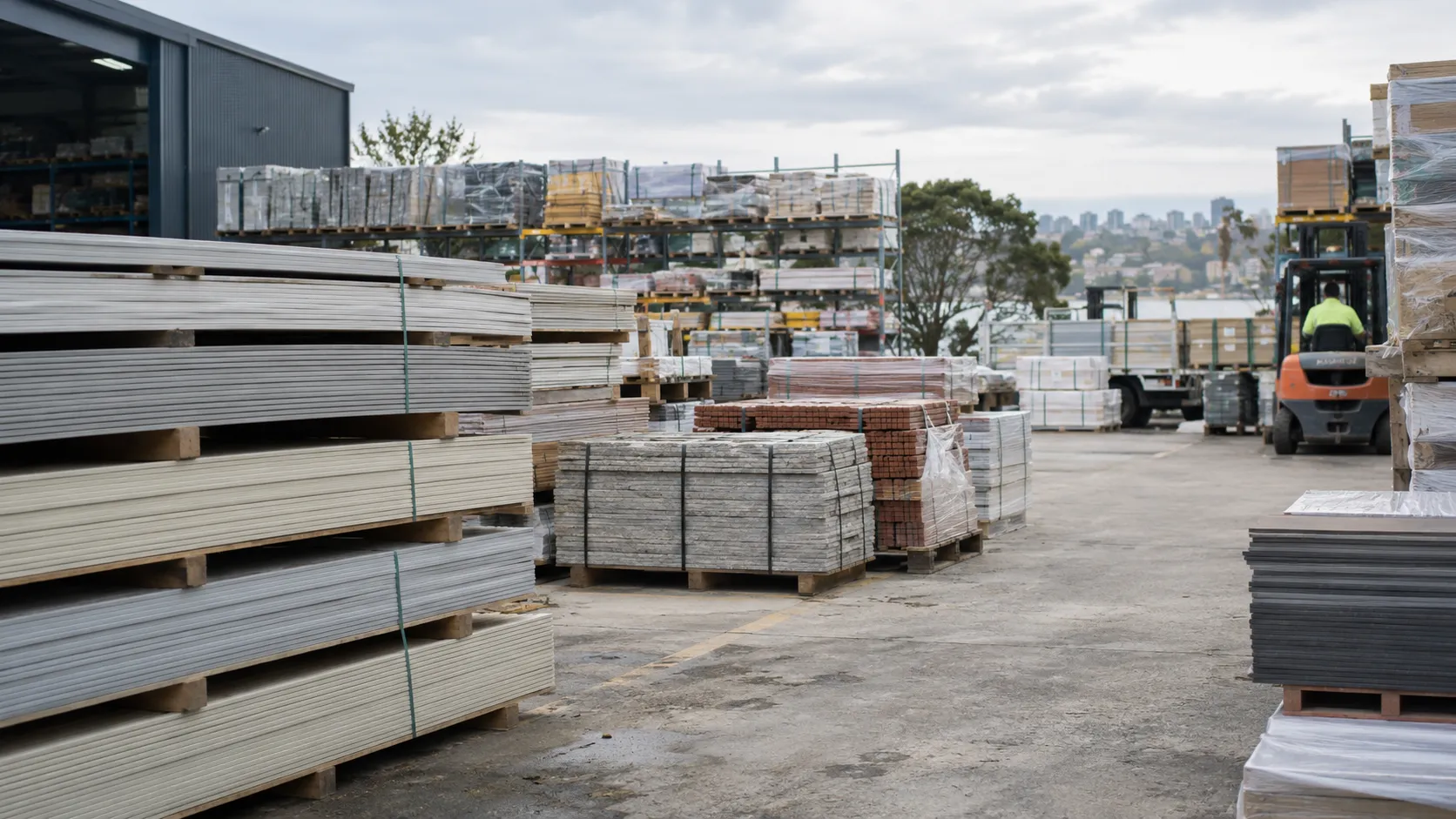 A building-materials yard with stacked boards, bricks and pavers on pallets, and a forklift working in the distance