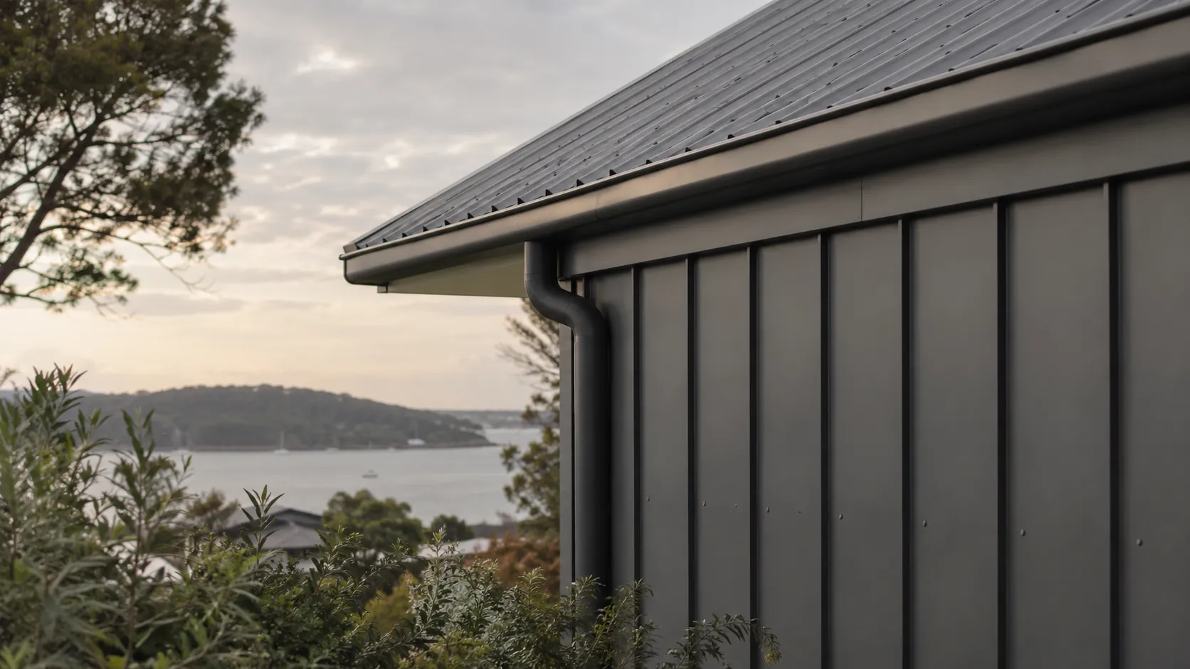 A dark steel roof edge with gutter, downpipe and vertical metal cladding overlooking a bay