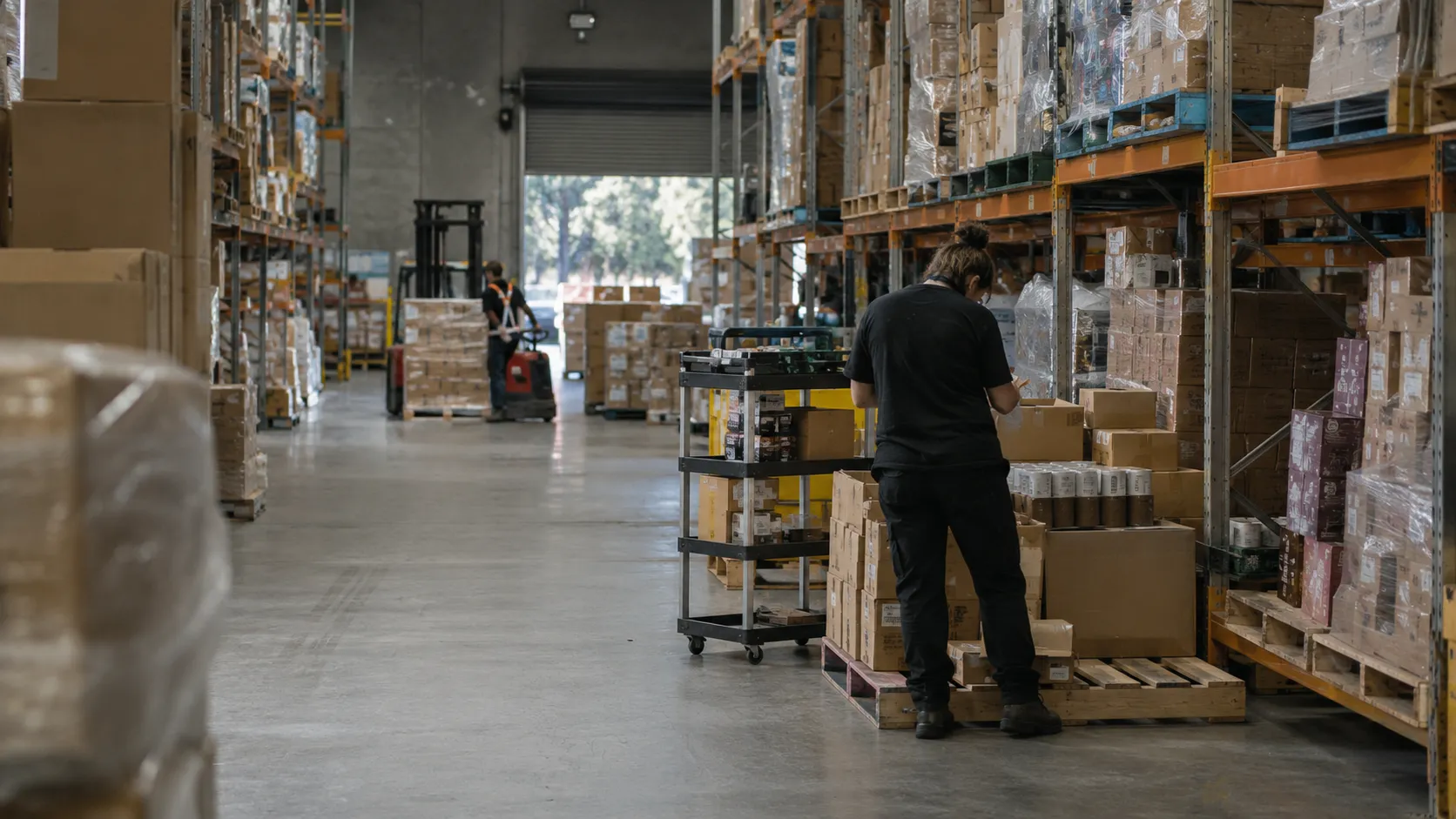 A warehouse worker checking stock on a cart between tall pallet racking, with a forklift operating near the loading door
