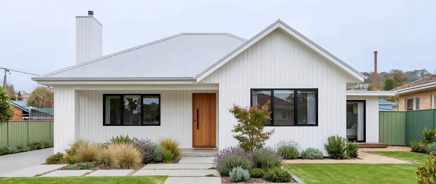 A single-storey house re-clad in white vertical cladding with a pale metal roof, black window frames, a timber front door and a new garden