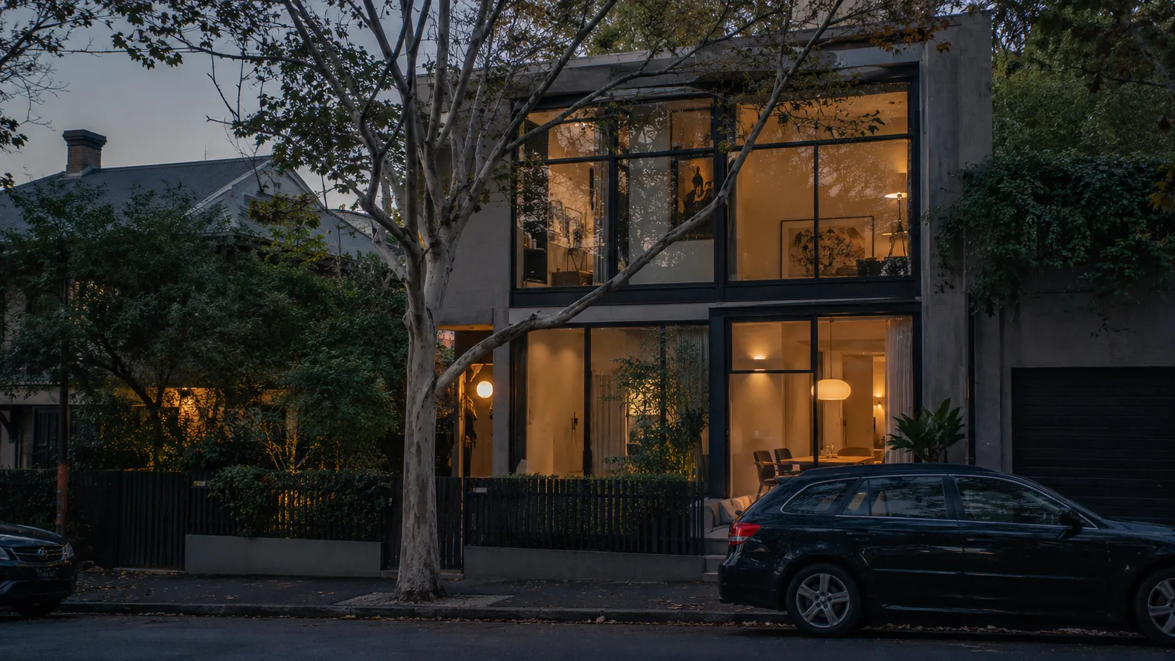 A modern townhouse at dusk with large black-framed windows glowing with warm interior light, a tree and a parked car