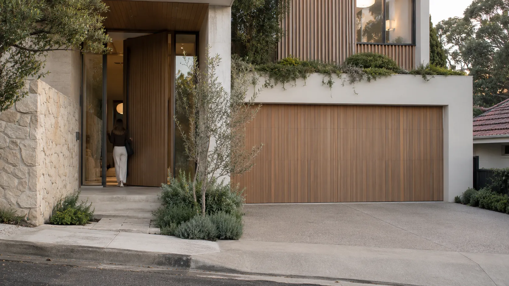 A house entrance with a tall timber pivot door, a matching timber garage door, a stone wall and planting