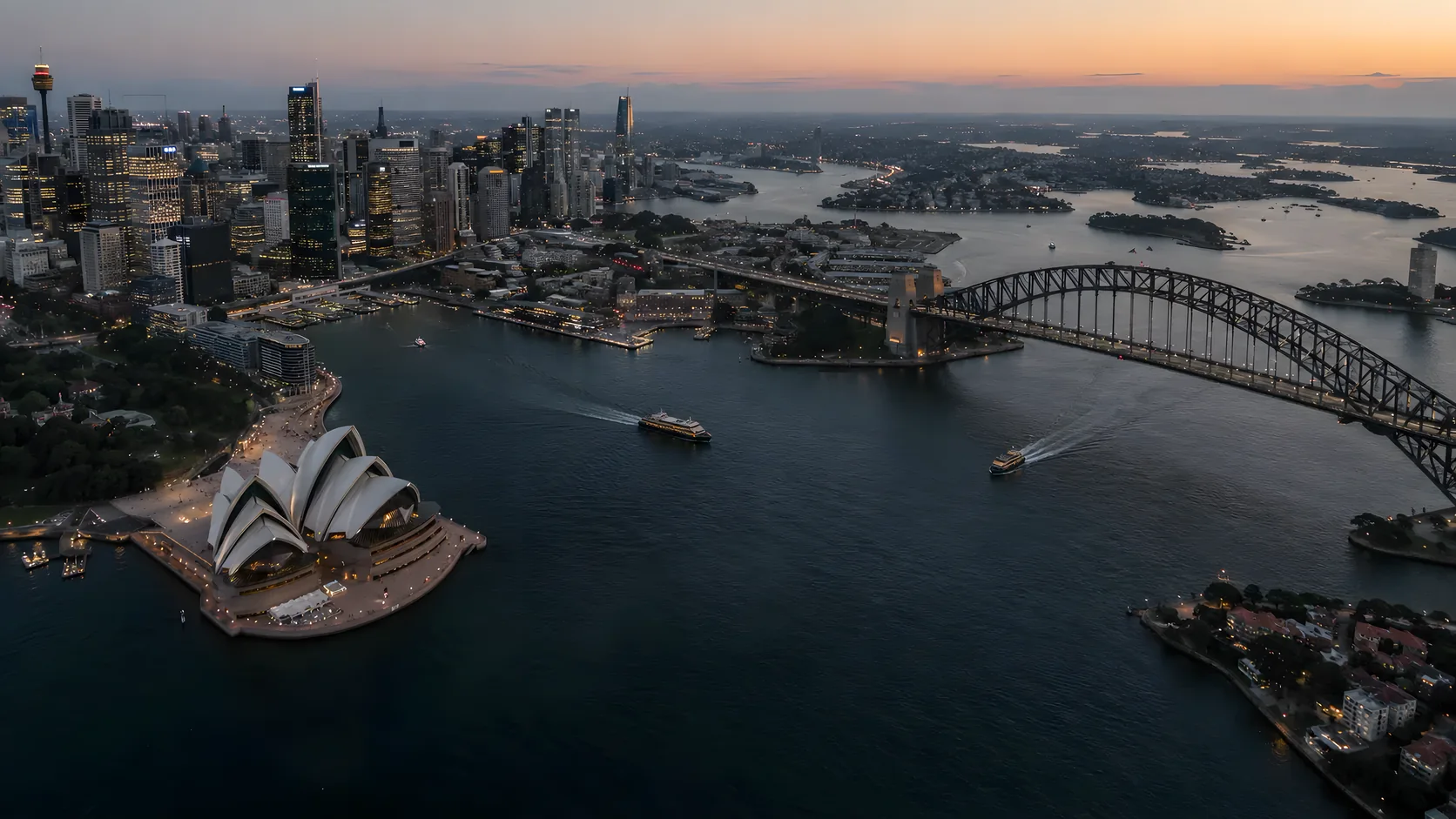 An aerial view of a harbour city at dusk, with the opera house, a steel arch bridge, ferries and a lit skyline